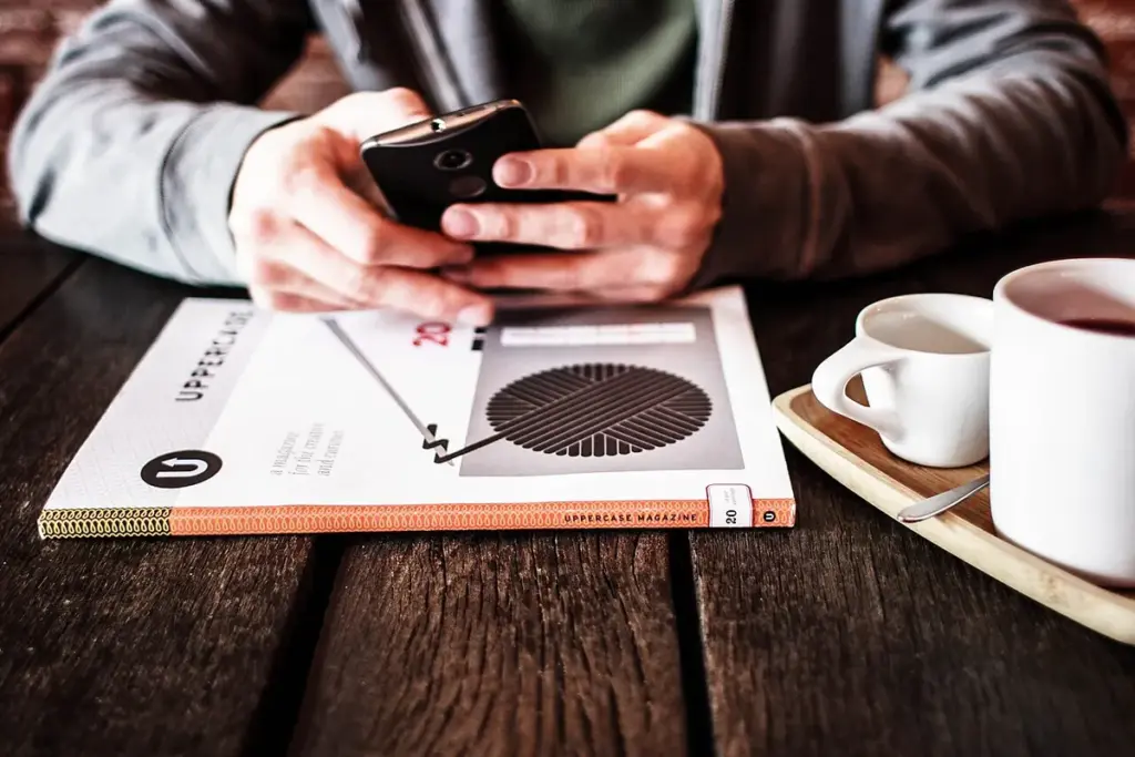 Pessoa segurando um celular sobre a mesa, ao lado de xícaras de café