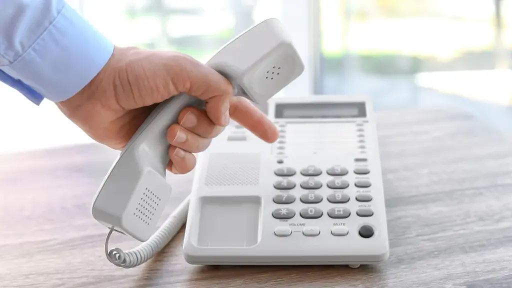 Mão segura o fone de um telefone fixo branco e pressiona uma tecla